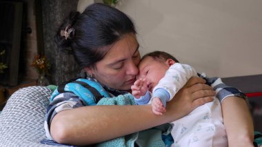 Anne kanepede otururken bebeğini nazikçe kucağına alıyor. Huzurlu ve sevgi dolu bir anne bakımı ve bağ kurma anı.