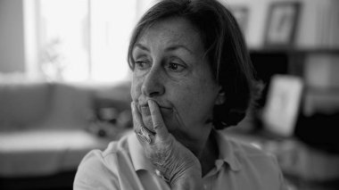 Düşünceli bir ifadeye sahip yaşlı bir kadın, elini çenesinde dinlendiriyor, ev ortamında, yumuşak ışıklandırmada, siyah ve beyaz bir anı yakalıyor.