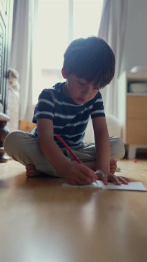 Çocuk yerde odaklanmış bir ifadeyle oturuyor, yardım alarak bir çizim etkinliğine katılıyor, konsantrasyon ve eğitim etkinliği gösteriyor