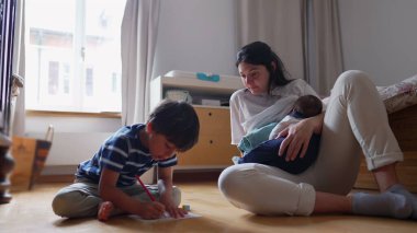 Anne, kucağında bebekle, bir ev okulu çizimi sırasında oğluna destek oluyor. Çok yönlü bir aile anı yakalıyor. Öğrenme, yaratıcılık ve beslenme.