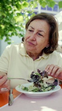 Yaşlı bir kadın açık hava kafede taze salata yiyor, canlı ve güneşli bir ortamda sağlıklı bir yemeğin tadını çıkarıyor, yaşlılar için sağlıklı ve dengeli bir diyet olduğunu vurguluyor. 