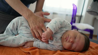 Ortopedist, çocukluğunda vücut ağrılarını hafifletmek için turuncu havluyla yeni doğan bebeğin pozisyonunu ayarlıyor, bebek rahat bir tulum giyiyor, dikkatli ve profesyonel yaklaşımı gösteriyor.