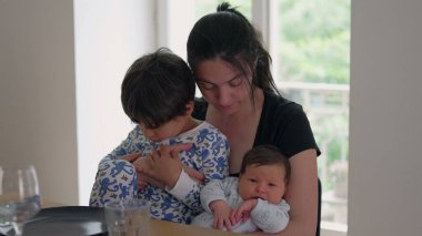 Mother holding her baby and young child, sitting at a table, highlighting family bonding moments. mother looks down lovingly while the children are in cozy pajamas clipart