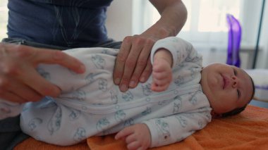 Ortopedist, yeni doğan bebeğin vücudunu turuncu bir havluyla düzelterek vücut ağrılarını hafifletiyor. Bebek esniyor ve uykuya dalıyor.
