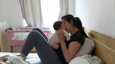 Anne bebeğini kucağında tutarken yatakta uzanıyor arka plandaki pencereden gün ışığıyla şefkatli bir öpücük paylaşıyor. Yumuşak ışık perdeleri ve pembe bebek beşiği olan rahat bir ev ortamı