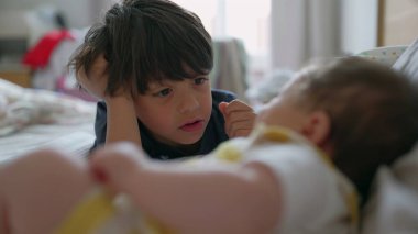 Büyük kardeş yatakta yatar, küçük kardeşlerine dikkatle bakar. Sessiz an, sıcak ve samimi bir ortamda ikisinin arasındaki bağ ve sevgiyi yansıtır.