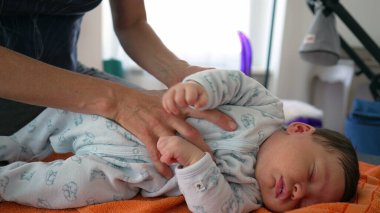 Ortopedist, çocukluk dönemindeki vücut ağrılarını hafifletmek için yeni doğan bebeği turuncu bir havluya nazikçe yerleştiriyor. Bebek bebek tulum giyer. Nazik ve profesyonel bakımı vurgular.