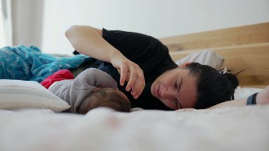 Anne bir yatakta uzanıyor, bebek dinlenirken sevgi dolu bir şekilde bebeğinin başına dokunuyor, sıcak ve davetkar bir yuva ortamında sakin ve besleyici bir etkileşimi temsil ediyor.