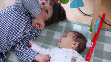 Küçük bir çocuk kareli bir oyun minderinde kardeşiyle sevgi dolu bir şekilde etkileşime girerken sevgi dolu aile ilişkileri ve değerli çocukluk anıları sergiliyor.