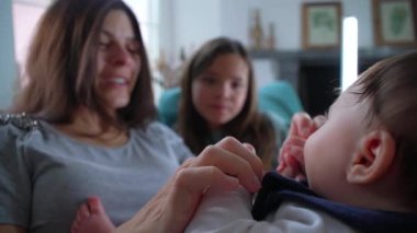 Bebeğin kucağına alınıp anne ve ablasına bakması sıcak bir aile anını her iki aile üyesinin de nazik etkileşimi ve ilgisiyle gösteriyor.