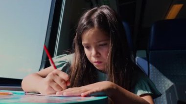 Genç kız yoğun bir şekilde trenle seyahat ederken kırmızı bir kalemle çizim yapmaya odaklanmış, pencerenin yanında oturmuş ve konsantrasyonlu bir masada çalışıyor.