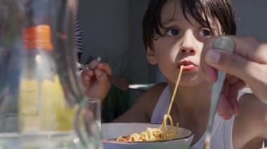 Genç çocuk spagetti yerken uzun bir erişteyi şapırdatarak yiyor, makarnayı çatalla çeviriyor ve gün boyunca dışarıda bir masada yemeğin tadını çıkarıyor.