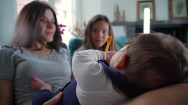 Anne ve kızı bir kanepede oturuyor, bir bebeği kucaklıyorlar ve nazikçe etkileşime geçiyorlar, sakin ve şefkatli bir aile anına odaklanıyorlar.