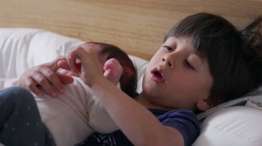Küçük çocuk yeni doğan kardeşini yatakta nazikçe tutuyor, sakin bir ifadeyle kameraya bakıyor, tatlı ve koruyucu bir kardeş anı yakalıyor.