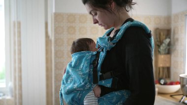 Anne bir mutfakta bebeğiyle birlikte duruyor, bebeği okşuyor, bir anlık şefkat ve bakım tasvir ediyor rahat ve şefkatli bir ev ortamında, aile yaşam tarzında.