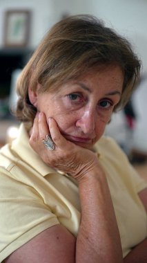 Düşünceli bir yüz ifadesiyle yaşlı bir kadın, yüzünü eline dayamış, bir anlık tefekkür ve sessiz bir yansıma tasvir ediyor.