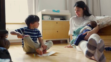 Anne oğlunu evde çizim yaparken izliyor, kucağında bebek, sıcak bir ev ortamında aile dinamiğini besliyor.
