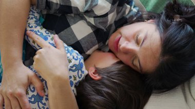 Anne ve çocuk kanepede birlikte uyukluyor, sıcak bir kucaklaşmayı paylaşıyor ve huzurlu bir ev ortamında ailevi sevgi ve konfor anı yakalıyor.