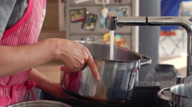Elleri mutfak musluğundan gelen suyla metal bir tencereyi dolduruyor. Sahne su akıntısına ve tencereye odaklanıyor. Mutfak ortamında yemek hazırlama aşamasında bir adım.