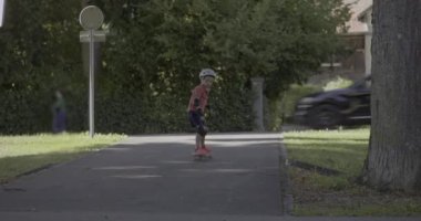 Genç bir çocuk parkta kaykay yapıyor, koruyucu giysi ve kask takıyor, yeşil ve kentsel unsurlarla çevrili, odaklı bir ifadeyle dengesini koruyor.