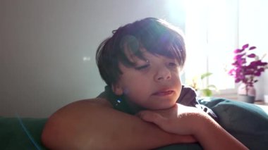 Genç çocuk burnunu ovalarken başını kollarının üzerinde dinlendiriyor. Güneş ışığı arka planda parlıyor ve içeride sakin ve yansıtıcı bir an yaratıyor..