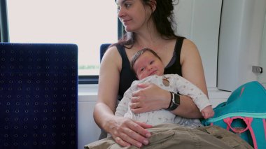 Anne bebeği trende tutuyor, hem mutlu görünüyor, hem de huzurlu bir aile seyahati, annelik bakımını, bağ kurmayı ve ortak bir yolculuğun huzurunu vurguluyor.