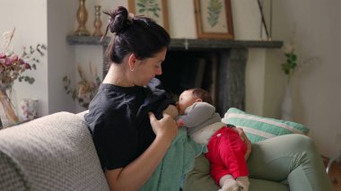 Bir kadın bebeğini kanepenin üzerinde emziriyor, arka planda şömine ve dekoratif öğeler var, sıcak, sade bir ortamda sessiz bir bağ kuruyor.
