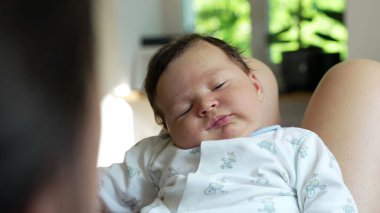 Ebeveyninin kucağında dinlenen bir bebeğin yakın çekimi, merak ve merakla bakıyor, şirin bir tulum giymiş, çocukluk yıllarının masumiyetini ve çekiciliğini vurguluyor.