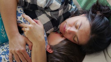 Anne ve çocuk kanepede birlikte uyukluyor, sıcak bir kucaklaşmayı paylaşıyor ve huzurlu bir ev ortamında ailevi sevgi ve konfor anı yakalıyor.