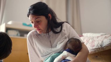 Anne yerde otururken bebeğini emziriyor, sıcak bir ev ortamında anne bakımı ve yakın ilişki anlarını vurguluyor.