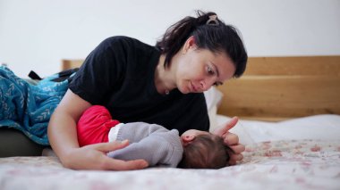 Anne bir yatakta uzanıyor, yatakta dinlenen bebeğini nazikçe okşuyor, basit, sakin bir ortamda duygusal ve samimi bir yakınlaşma ve sevgi anı yaşıyor.