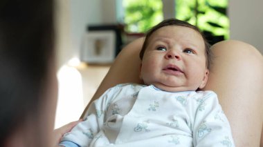 Ebeveyninin kucağında dinlenen bir bebeğin yakın çekimi, merak ve merakla bakıyor, şirin bir tulum giymiş, çocukluk yıllarının masumiyetini ve çekiciliğini vurguluyor.