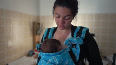 Anne, mutfakta bebek taşıyıcısı taşırken bebeğine bakıyor. Aile hayatının şefkatli ve samimi anlarını vurguluyor.