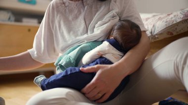 Anne yerde otururken bebeğini emziriyor, sıcak bir ev ortamında anne bakımı ve yakın ilişki anlarını vurguluyor.