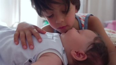 Büyük çocuk yatağın üzerinde bebeğe yakın yatıyor, sakin ve sevgi dolu bir bağ gösterirken başını dinlendiriyor. Bebekle kardeşler arasında kameraya doğru bakan kapalı bir ortam.