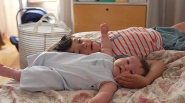 Büyük çocuk yatağın üzerinde bir bebeğin yanında uzanıyor, sakin bir iç mekan ortamında dinleniyor. Bebek gülümserken büyük çocuk bakar, sessiz bir kardeş yakınlaşması anı