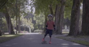 Korumalı giysiler giymiş, elinde kaykayla parkın patikasında duran genç bir çocuk etrafı ağaçlarla çevrili olan mesafeye bakıyor.