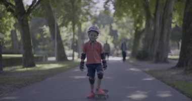 Korumalı giysiler giyen genç çocuk park yolunda kaykay yapmaya kalkışıyor, kollarını denge için uzatıyor, gölgeli doğal ortamda kararlılık ve çaba gösteriyor.