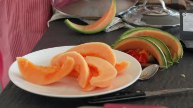 Taze kavun dilimleri mutfak tezgahındaki beyaz tabakta sıralanmış. Meyvenin canlı turuncu rengi yeşil kabukla çelişiyor ve görsel olarak çekici bir sunum oluşturuyor.