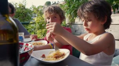 İki çocuk açık hava yemeğinden zevk alıyor, tabağında çatalla spagetti karıştıran çocuğa odaklanıyor. Arka planda yeşillik olan sıradan bir aile yemeği.