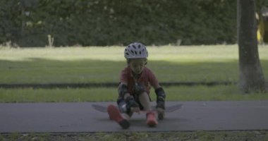 Genç bir çocuk, kaskı ve koruyucu giysileri ile parkta oturuyor, bacaklarının arasında kaykay tutuyor, sakin ve odaklanmış bir şekilde bir sonraki hamlesine hazırlanıyor.