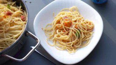 Beyaz bir tabakta servis edilen spagetti porsiyonu taze pişmiş bir makarna tavasının yanında. Ortam rahat, aile ve arkadaşlarla rahat bir açık hava yemeği için mükemmel.