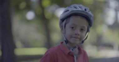 Güneşli bir günde parkın doğal yeşiliyle çevrili sakin ve odaklanmış bir ifadeyle ileriye bakan kask takan genç bir çocuğun yakın çekimi.