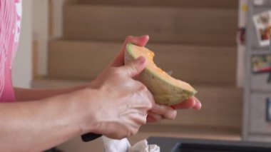 Mutfak tezgahında taze kavun dilimleri kesen eller, canlı meyvenin servis için hazırlandığını gösteriyor. Mutfak düzeninde çeşitli mutfak aletleri ve çizgili bir mutfak havlusu var.