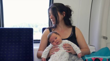 Anne, yeni doğmuş bir bebeği kollarında bir trende, pencerenin yanında tutuyor. Yolculuğun hassas anlarını ve anne ile çocuk arasındaki bağı vurguluyor.