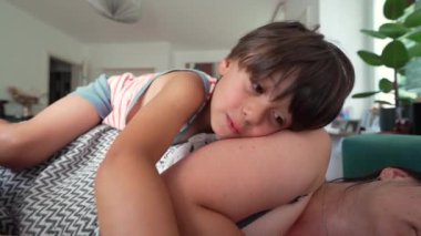 Genç çocuk, annesi uyurken başını kanepede uzanan bir bebeğin yanında dinlendiriyor. Kardeşlerin sevgisini, annelerin sıcaklığını yakalayan huzurlu bir aile anı.