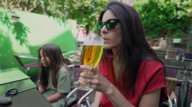 Genç kızı yakınlarda otururken, annesi açık bir kafede serinletici bir içki yudumluyor. Aile gezisinin rahatlatıcı, sıradan havası.