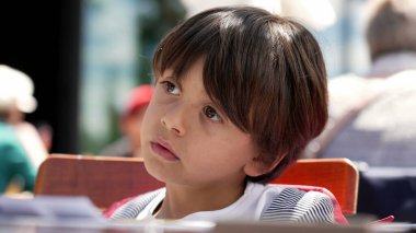 Düşünceli bir ifadeyle, açık bir kafe masasında oturan genç bir çocuğun sosyal ve canlı bir ortamda çocukluk merakını ve düşüncelerini vurguluyor.