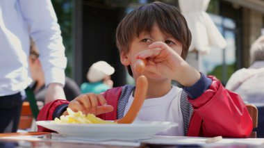 Kırmızı ceketli çocuk, açık hava restoranında sosis ve patates kızartması yiyor. Sunucu güneşli bir günde arka planda tabak getiriyor.