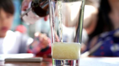 Açık hava bir kafede biranın bardağa doldurulduğu yakın plan bir sosyal ortamın bulanık arka planı, soğuk bir içecekten keyif almanın ferahlatıcı doğasını vurguluyor.